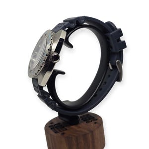 May include: A stainless steel wristwatch with a dark blue rubber strap. The watch has a round face with a light blue bezel and a black dial. The watch is displayed on a dark blue watch stand with a wooden base.