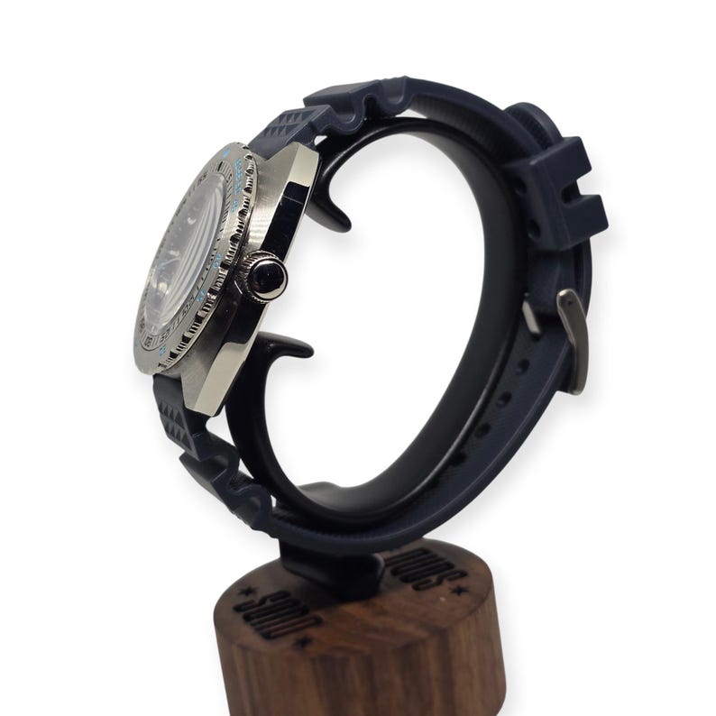 May include: A stainless steel wristwatch with a dark blue rubber strap. The watch has a round face with a light blue bezel and a black dial. The watch is displayed on a dark blue watch stand with a wooden base.