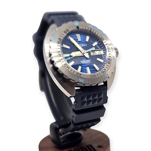May include: A stainless steel Seiko automatic watch with a blue wave-patterned dial and a black rubber strap. The watch features a date display and a rotating bezel with blue accents. The watch is displayed on a black stand.