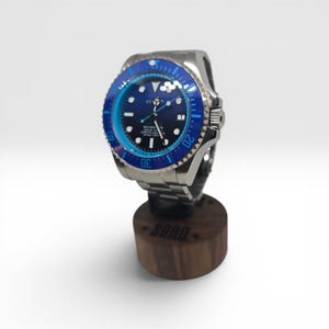 May include: A stainless steel wristwatch with a dark blue dial and a rotating blue bezel featuring white markers. The watch face displays the text SEA-DWELLER, 12800FT=3900M, and LIMITED EDITION. It is mounted on a cylindrical dark wood display stand.