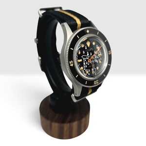 May include: A Seiko automatic wristwatch with a black dial featuring a white grid pattern, luminous markers, and an orange second hand. The watch has a black rotating bezel with gold numerals and is fitted with a black and gold striped fabric strap.