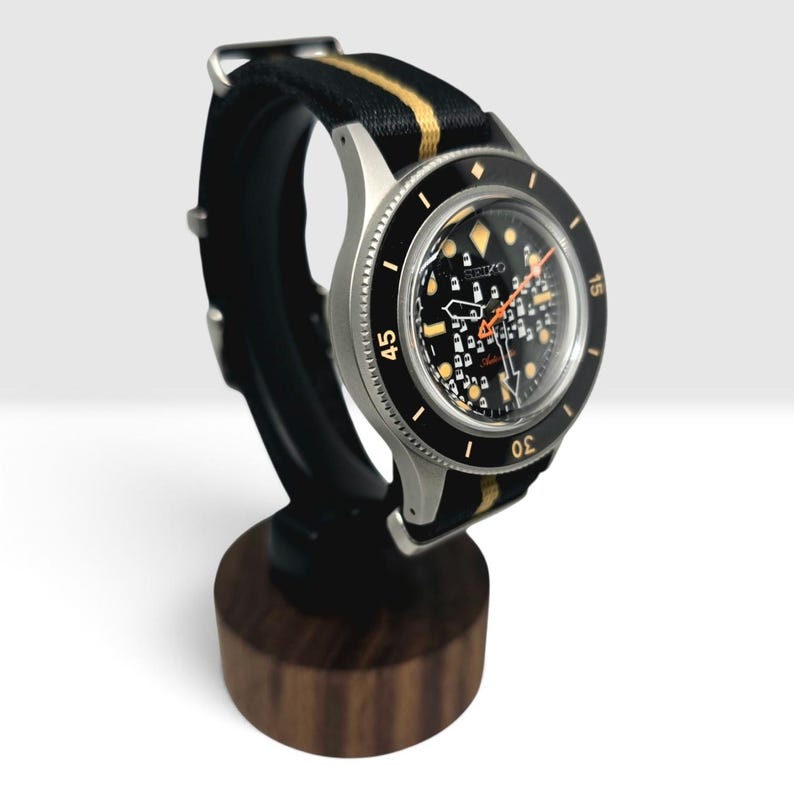 May include: A Seiko automatic wristwatch with a black dial featuring a white grid pattern, luminous markers, and an orange second hand. The watch has a black rotating bezel with gold numerals and is fitted with a black and gold striped fabric strap.