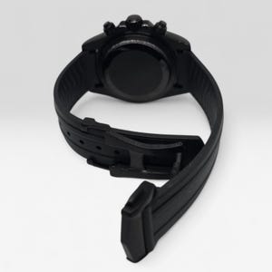 May include: A matte black wristwatch featuring a circular case with three side buttons and a textured silicone strap. The watch includes a black buckle closure system and a smooth, solid black case back, presented from a rear-facing perspective.