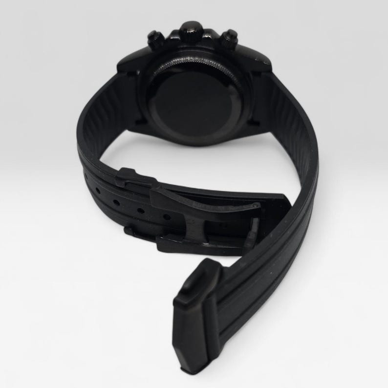 May include: A matte black wristwatch featuring a circular case with three side buttons and a textured silicone strap. The watch includes a black buckle closure system and a smooth, solid black case back, presented from a rear-facing perspective.