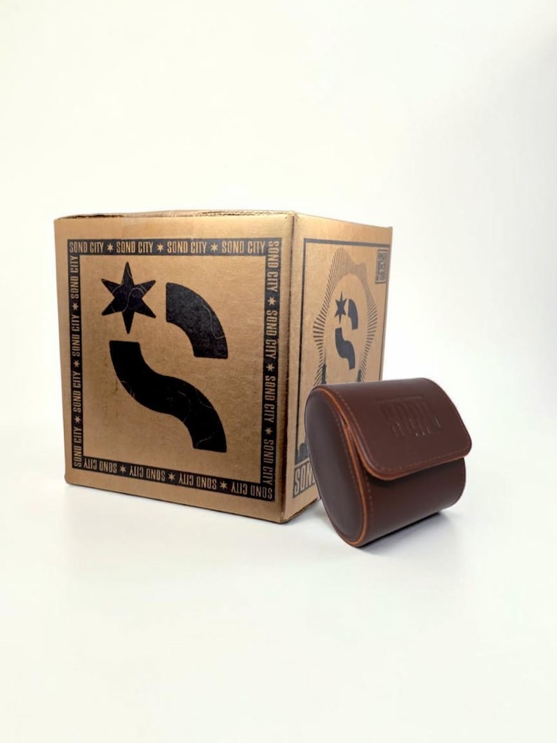 Puede incluir: Un estuche cil&iacute;ndrico para reloj de color marr&oacute;n con cierre de solapa se encuentra junto a una caja de cart&oacute;n cuadrada. La caja presenta gr&aacute;ficos impresos en negro que incluyen una estrella, una forma curva y el texto SOND CITY repetido.