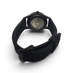 May include: A matte black wristwatch featuring a flexible silicone strap and a circular case. The back of the watch is transparent, revealing the intricate metallic mechanical movement components inside the casing, including a silver-toned rotor.