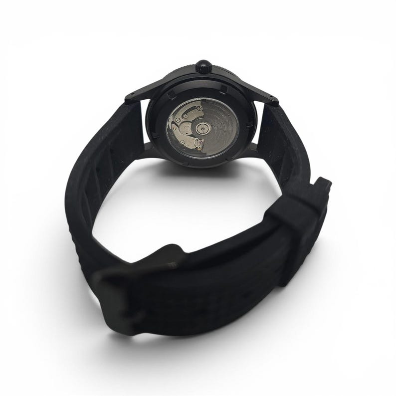 May include: A matte black wristwatch featuring a flexible silicone strap and a circular case. The back of the watch is transparent, revealing the intricate metallic mechanical movement components inside the casing, including a silver-toned rotor.