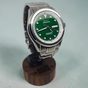 May include: A silver-tone stainless steel wristwatch with a textured green dial, featuring the text SEIKO AUTOMATIC and OCEAN STAR DIVER'S 200M. The watch has a day-date window at the three o'clock position, luminous hands, and a metal link bracelet.