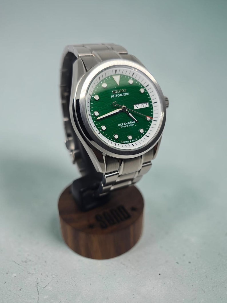 May include: A silver-tone stainless steel wristwatch with a textured green dial, featuring the text SEIKO AUTOMATIC and OCEAN STAR DIVER'S 200M. The watch has a day-date window at the three o'clock position, luminous hands, and a metal link bracelet.