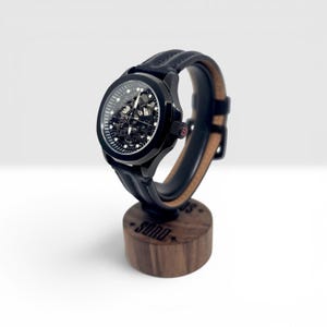 May include: A black skeleton watch with a visible mechanical movement, a round case, and a black leather strap with visible stitching. The watch is mounted on a cylindrical wooden stand featuring the text SONO engraved on the front surface.