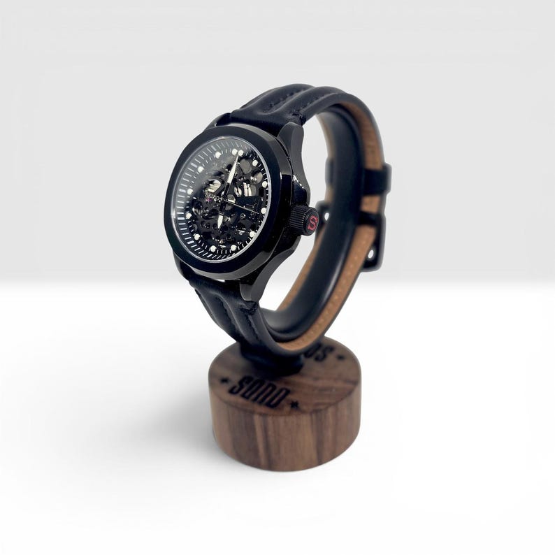 May include: A black skeleton watch with a visible mechanical movement, a round case, and a black leather strap with visible stitching. The watch is mounted on a cylindrical wooden stand featuring the text SONO engraved on the front surface.