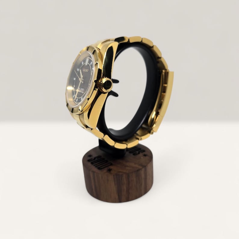 Op de afbeelding: Een goudkleurig polshorloge met een zwarte wijzerplaat en gouden wijzers wordt getoond op een zwarte standaard. Het horloge heeft een gouden band en een ronde wijzerplaat. De standaard is gemaakt van donker hout en heeft de inscriptie "SOUD".