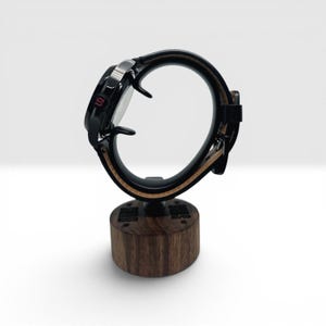May include: A black wristwatch with a dark leather strap featuring a light brown underside, mounted on a cylindrical wooden display stand. The watch case has a small red letter S on the side, and the wooden base is engraved with text and stars.