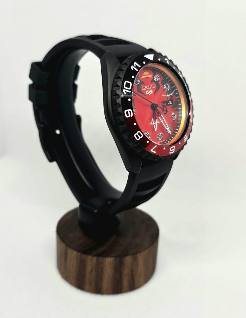 May include: A Seiko 5 Sports automatic watch with a black case and silicone strap. The dial is red with a straw hat icon at the twelve o'clock position, black indices, and a date window. The bezel is black and red with white numerals.