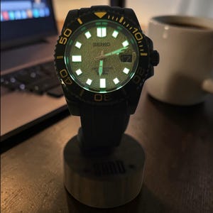 Pu&ograve; includere: Un orologio da polso automatico Seiko nero con quadrante testurizzato di colore chiaro, indici e lancette luminescenti. L'orologio presenta una lunetta girevole con accenti gialli, una finestrella per la data e un cinturino in gomma nera.