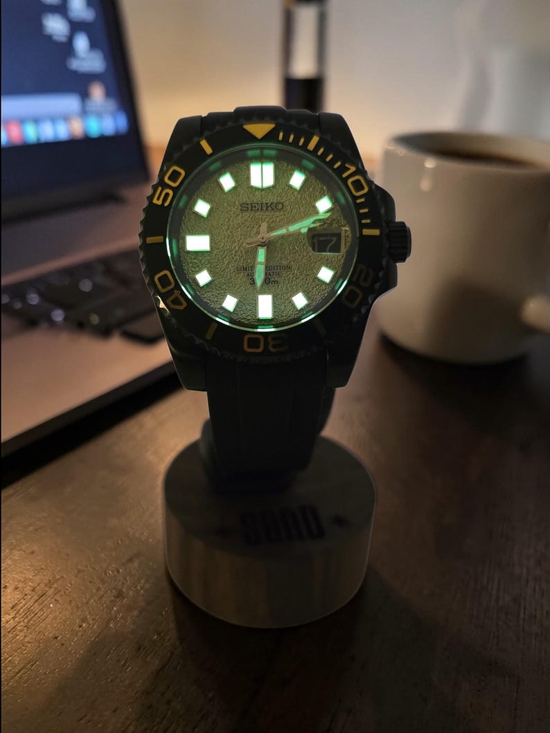 Pu&ograve; includere: Un orologio da polso automatico Seiko nero con quadrante testurizzato di colore chiaro, indici e lancette luminescenti. L'orologio presenta una lunetta girevole con accenti gialli, una finestrella per la data e un cinturino in gomma nera.