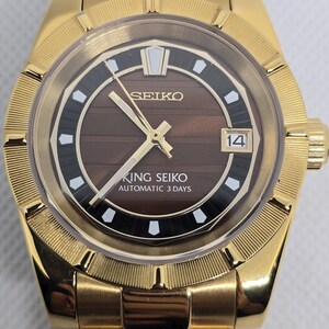 Op de afbeelding: Goudkleurig Seiko King Seiko automatisch polshorloge. Het horloge heeft een bruine wijzerplaat met gouden wijzers en markeringen, een datumweergave en de tekst "KING SEIKO AUTOMATIC 3 DAYS". Het horloge heeft een goudkleurige armband.