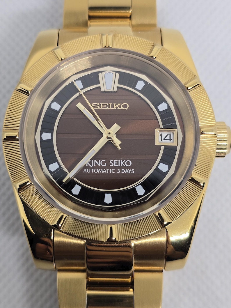 Op de afbeelding: Goudkleurig Seiko King Seiko automatisch polshorloge. Het horloge heeft een bruine wijzerplaat met gouden wijzers en markeringen, een datumweergave en de tekst "KING SEIKO AUTOMATIC 3 DAYS". Het horloge heeft een goudkleurige armband.