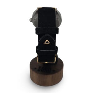 May include: A wristwatch with a black strap featuring a small triangular gold stitch detail, resting on a cylindrical wooden stand with the word SOHO printed on the front. The watch has a metallic casing and a gold-toned buckle on the strap.
