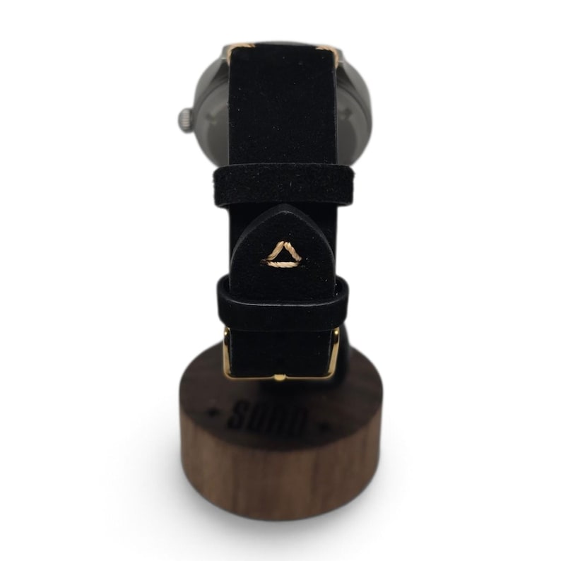 May include: A wristwatch with a black strap featuring a small triangular gold stitch detail, resting on a cylindrical wooden stand with the word SOHO printed on the front. The watch has a metallic casing and a gold-toned buckle on the strap.