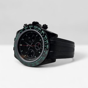 May include: A black chronograph wristwatch featuring a black dial with three sub-dials, a tachymeter bezel with green numerals, and a black rubber strap. The watch includes silver-toned hands with red accents and a date window at the three o'clock position.