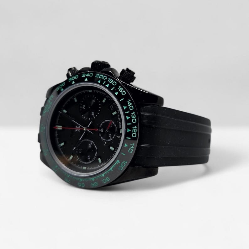 May include: A black chronograph wristwatch featuring a black dial with three sub-dials, a tachymeter bezel with green numerals, and a black rubber strap. The watch includes silver-toned hands with red accents and a date window at the three o'clock position.