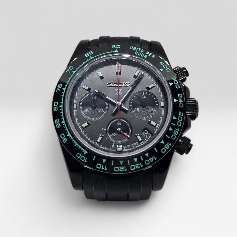 May include: A black Seiko chronograph watch with a dark grey dial, three sub-dials, and a tachymeter bezel featuring teal-colored numbers and the text UNITS PER HOUR. The watch includes a date window at the four o'clock position and silver hands.