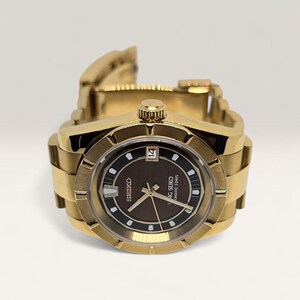 Op de afbeelding: Een goudkleurig polshorloge met een bruine wijzerplaat en een datumweergave. Het horloge heeft een getextureerde bezel en een gouden schakelarmband. De wijzerplaat toont de merknaam "Seiko" en de woorden "Automatic 3 Days".