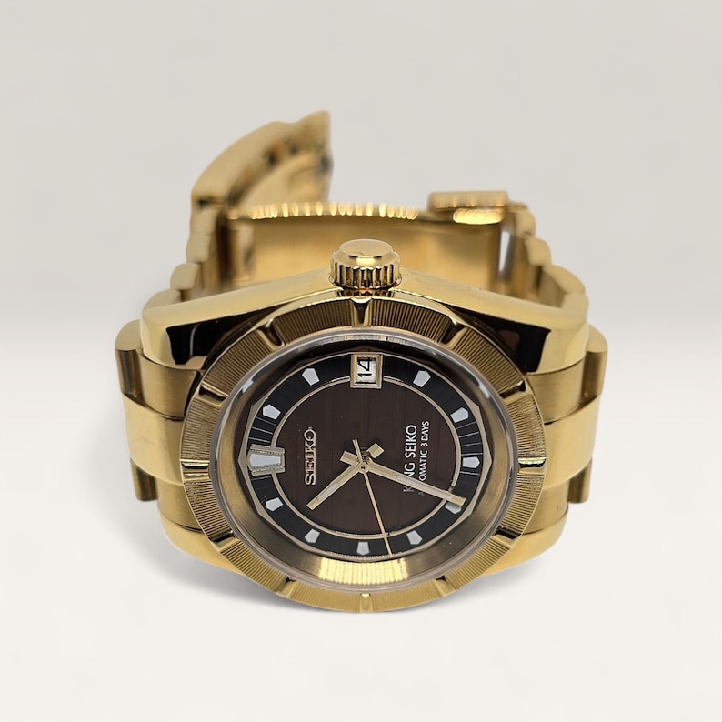 Op de afbeelding: Een goudkleurig polshorloge met een bruine wijzerplaat en een datumweergave. Het horloge heeft een getextureerde bezel en een gouden schakelarmband. De wijzerplaat toont de merknaam "Seiko" en de woorden "Automatic 3 Days".