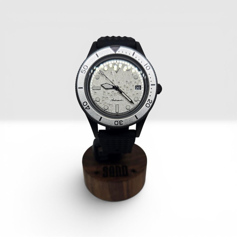 May include: An automatic wristwatch with a textured silver dial, black hands, and a date window at the three o'clock position. The watch features a silver rotating bezel with black numerals and a black case and strap, mounted on a wooden stand.