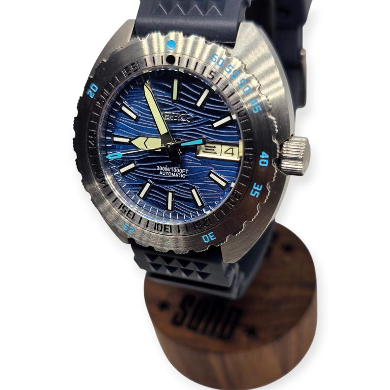 May include: A stainless steel wristwatch with a blue wave-patterned dial and a black rubber strap. The watch features a date display and luminous hands. The bezel has blue and white markings. The watch is displayed on a wooden stand.