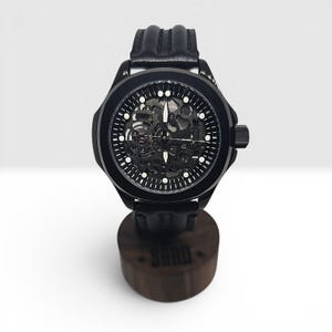 May include: A black wristwatch with a skeleton dial revealing internal mechanical components, white hour markers, and luminous hands. The watch features a black case and a textured black leather strap, resting on a cylindrical wooden stand.