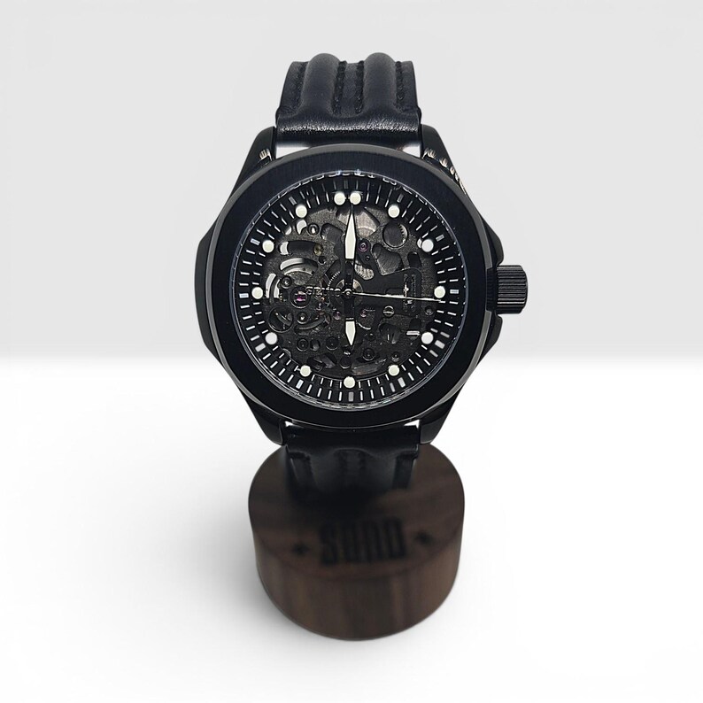 May include: A black wristwatch with a skeleton dial revealing internal mechanical components, white hour markers, and luminous hands. The watch features a black case and a textured black leather strap, resting on a cylindrical wooden stand.