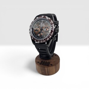 K&ouml;nnte beinhalten: Eine schwarze Seiko Chronographenuhr mit braunem Zifferblatt, drei Hilfszifferbl&auml;ttern und Tachymeterl&uuml;nette, montiert auf einem zylindrischen Holzst&auml;nder mit der Gravur SQND auf der Vorderseite. Die Uhr hat ein schwarzes Kautschukarmband.