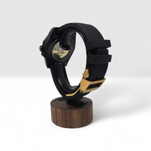 Pu&ograve; includere: Un orologio da polso nero con cinturino in silicone e fibbia dorata, montato su un supporto nero curvo sopra una base cilindrica in legno scuro. Il fondello dell'orologio &egrave; trasparente, rivelando il movimento meccanico e il rotore dorato.