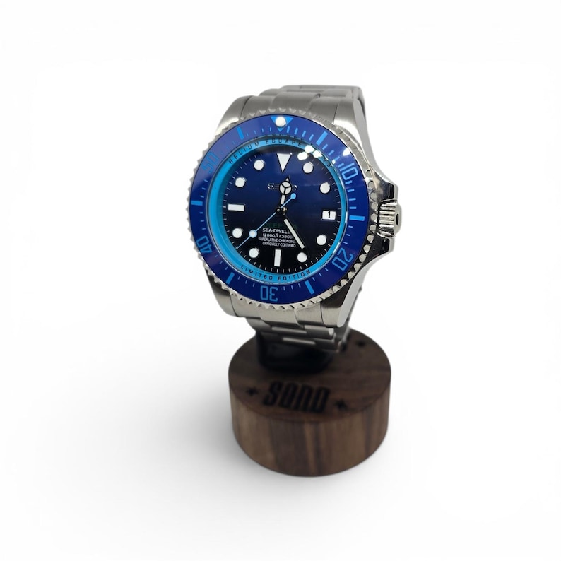 May include: A silver-tone stainless steel wristwatch with a dark blue dial and a rotating blue bezel. The watch face features white hour markers, a date window, and the text SEA-DWELLER. It is mounted on a cylindrical dark wood display stand.