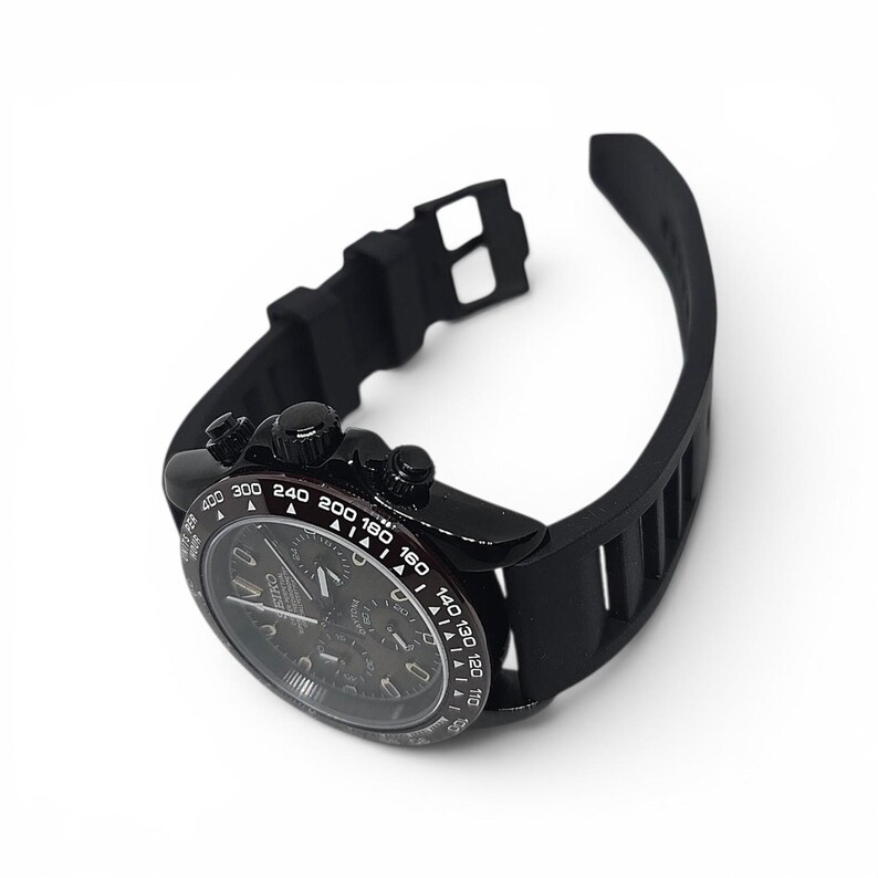 K&ouml;nnte beinhalten: Eine schwarze Chronographen-Armbanduhr mit rundem Geh&auml;use, Tachymeter-L&uuml;nette mit wei&szlig;en Markierungen und schwarzem Silikonarmband. Das Zifferblatt zeigt drei Hilfszifferbl&auml;tter, leuchtende Zeiger und den Markennamen Seiko in wei&szlig;er Schrift.