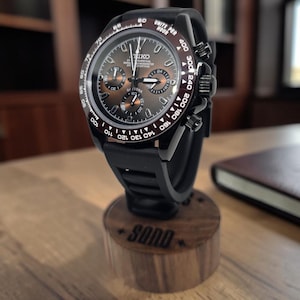 K&ouml;nnte beinhalten: Eine schwarze Chronographen-Armbanduhr mit dunkelbraunem Sonnenschliff-Zifferblatt, drei Hilfszifferbl&auml;ttern und Tachymeter-L&uuml;nette. Die Uhr hat ein schwarzes Kautschukarmband und steht auf einem zylindrischen Holzst&auml;nder mit der Gravur SOND.