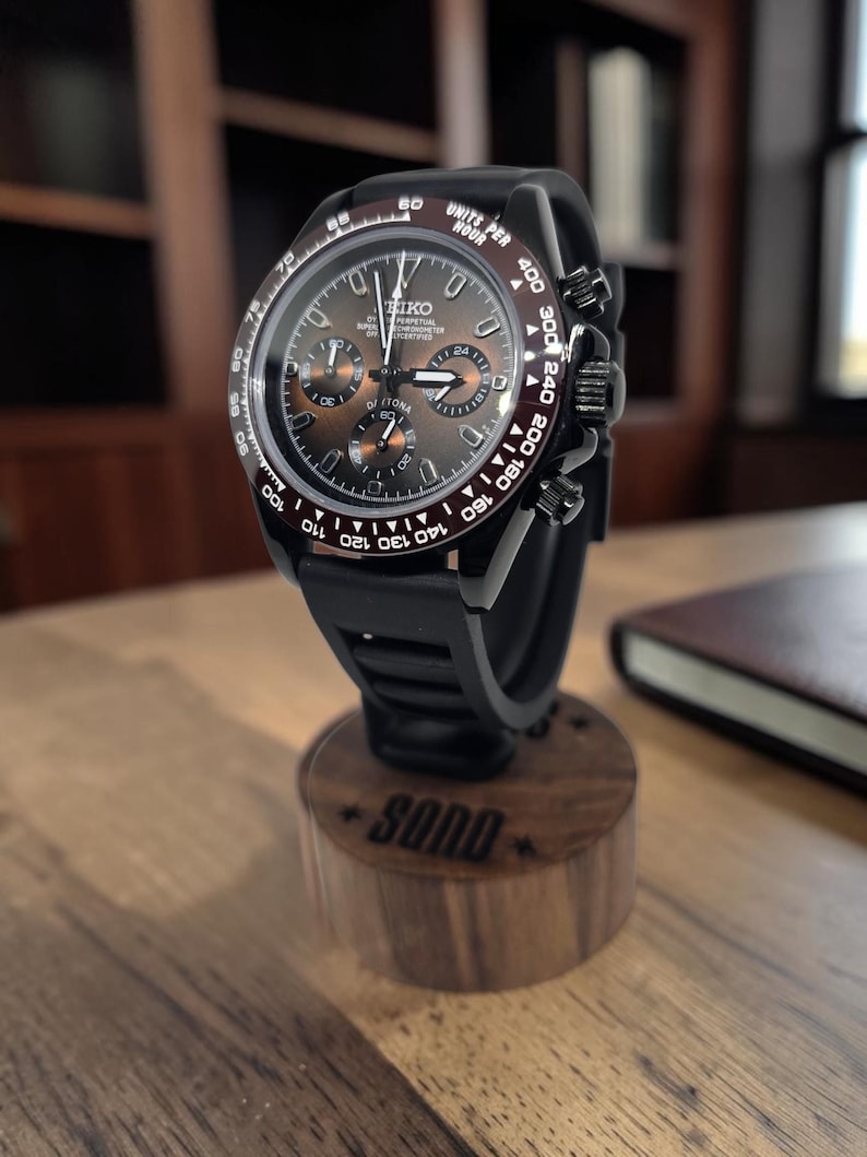 K&ouml;nnte beinhalten: Eine schwarze Chronographen-Armbanduhr mit dunkelbraunem Sonnenschliff-Zifferblatt, drei Hilfszifferbl&auml;ttern und Tachymeter-L&uuml;nette. Die Uhr hat ein schwarzes Kautschukarmband und steht auf einem zylindrischen Holzst&auml;nder mit der Gravur SOND.