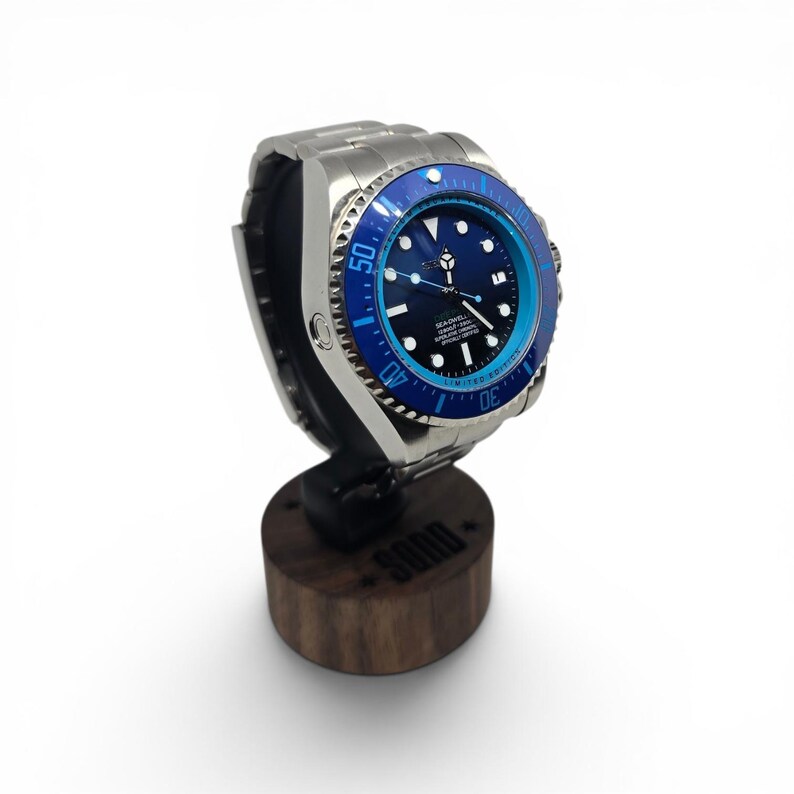 May include: A stainless steel wristwatch with a black dial and a rotating blue bezel featuring white numerals. The watch has a metal link bracelet and is displayed on a cylindrical dark wood stand with the word SODO engraved on the front.