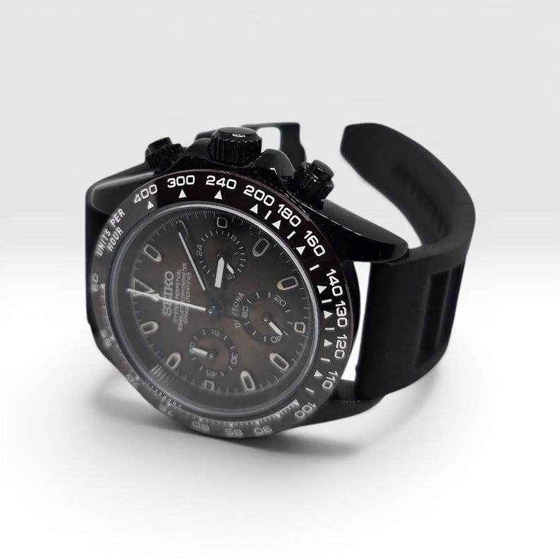 K&ouml;nnte beinhalten: Eine schwarze Chronographen-Armbanduhr mit dunklem Zifferblatt, drei Hilfszifferbl&auml;ttern, einer Tachymeterskala auf der L&uuml;nette mit wei&szlig;en Markierungen und einem schwarzen Silikonarmband. Das Zifferblatt zeigt das Seiko-Logo und Units Per Hour.