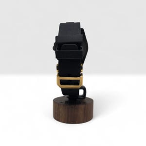 Pu&ograve; includere: Un cinturino per orologio in silicone nero con fibbia in metallo color oro, esposto verticalmente su un supporto cilindrico in legno. Il cinturino presenta una finitura opaca liscia con un passante rettangolare e una chiusura ad ardiglione.
