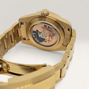 Op de afbeelding: Goudkleurig polshorloge met een gedetailleerde armband. De wijzerplaat heeft een ontwerp met twee draken, een teal en een bruine, en andere symbolen. Het horloge wordt van achteren getoond, waarbij de armband gedeeltelijk zichtbaar is.