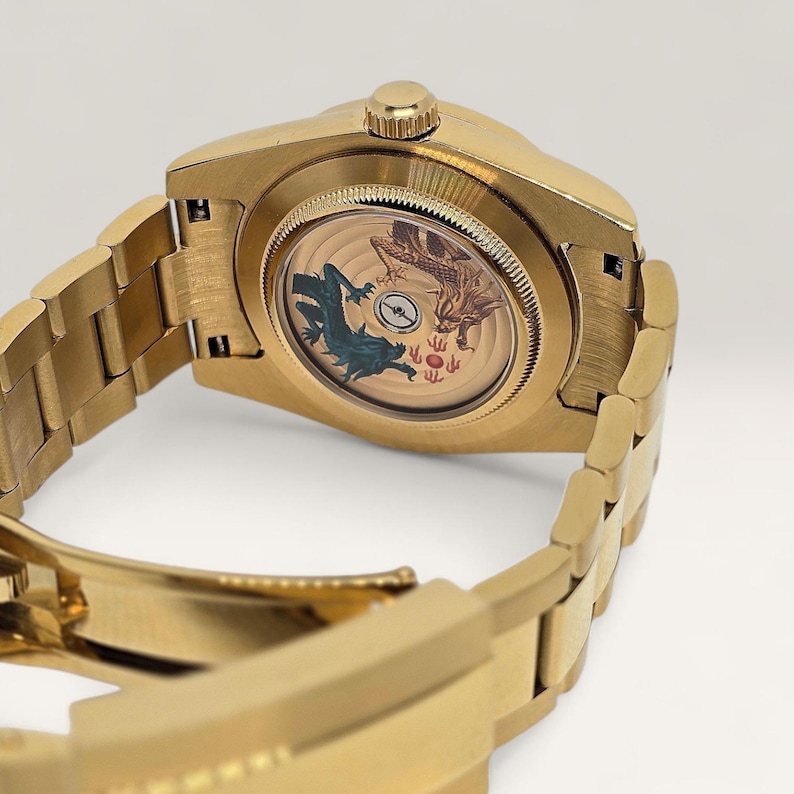 Op de afbeelding: Goudkleurig polshorloge met een gedetailleerde armband. De wijzerplaat heeft een ontwerp met twee draken, een teal en een bruine, en andere symbolen. Het horloge wordt van achteren getoond, waarbij de armband gedeeltelijk zichtbaar is.