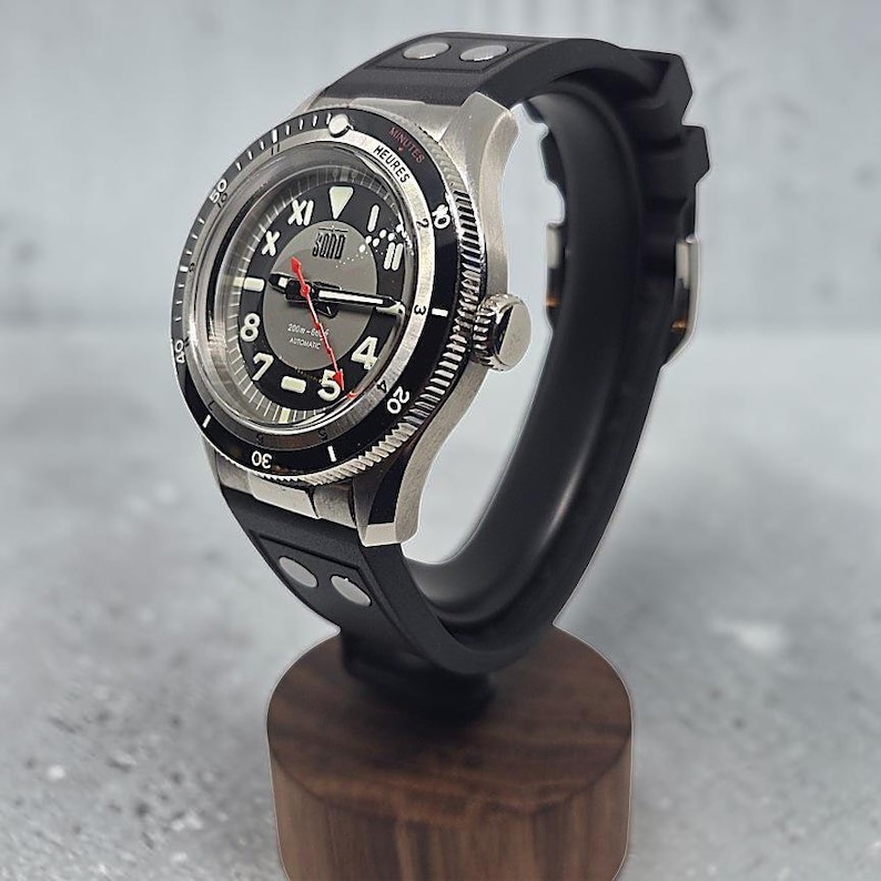 Puede incluir: Un reloj de pulsera autom&aacute;tico de acero inoxidable con correa de caucho negra y dos remaches plateados. La esfera es negra y gris con n&uacute;meros blancos, segundero rojo y un bisel giratorio negro marcado con minutos y la palabra HEURES.