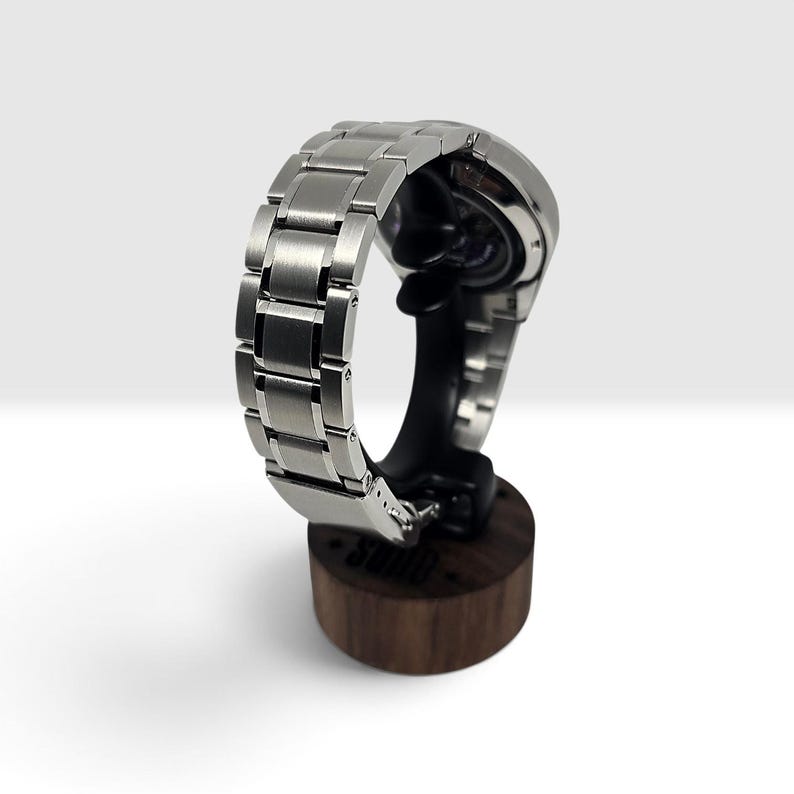 May include: A silver-toned metal wristwatch with a brushed link bracelet, displayed on a dark brown cylindrical wooden stand. The watch case features a circular design with a visible mechanical movement on the back, resting securely on the stand.