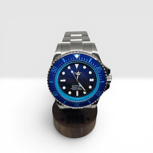 May include: A stainless steel wristwatch with a dark blue dial and a rotating bezel featuring a blue and light blue color scheme. The watch has luminous hour markers, silver hands, and a metal link bracelet, displayed on a cylindrical stand.