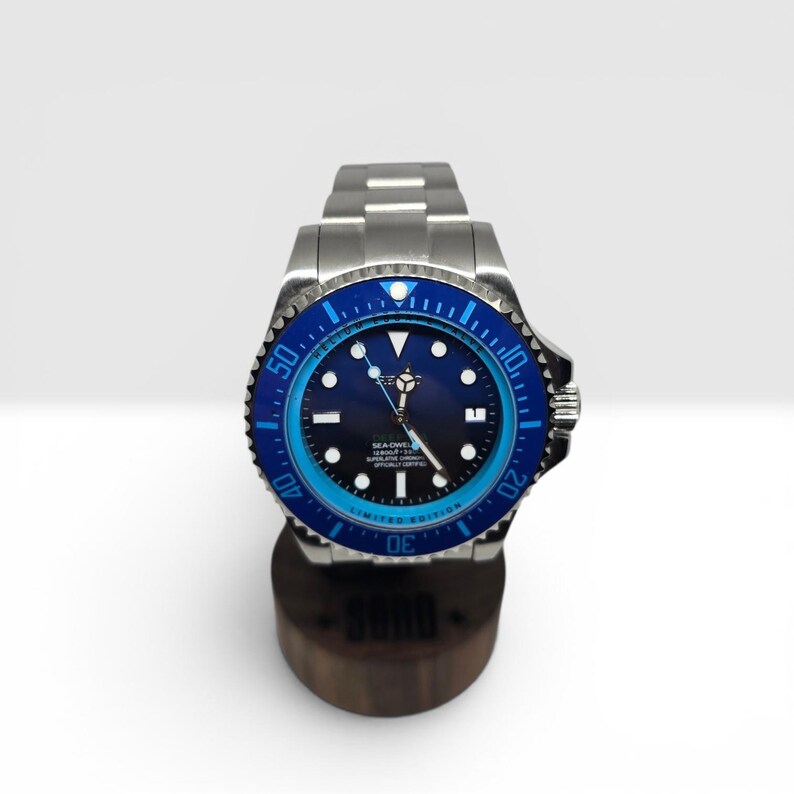 May include: A stainless steel wristwatch with a dark blue dial and a rotating bezel featuring a blue and light blue color scheme. The watch has luminous hour markers, silver hands, and a metal link bracelet, displayed on a cylindrical stand.