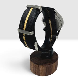 May include: A wristwatch featuring a black nylon strap with a central tan stripe and silver-toned metal hardware. The watch case is circular with a metallic finish, and the entire piece is displayed on a dark wood cylindrical stand.