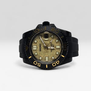 Pu&ograve; includere: Un orologio da polso Seiko con quadrante dorato testurizzato, indici quadrati luminescenti, ghiera girevole nera con numeri dorati e cinturino in silicone nero. L'orologio presenta una finestrella per la data a ore tre e lancette argentate.